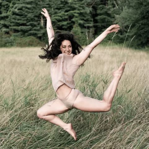 A person in mid-air leaping above a grassy field with evergreen trees.